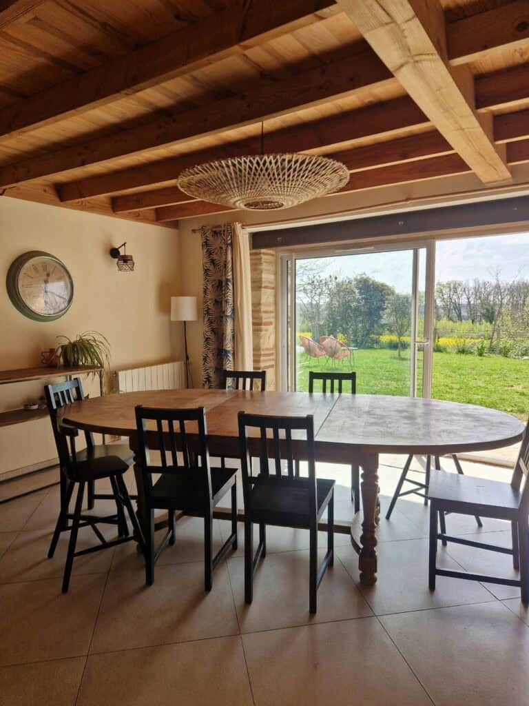 Une salle à manger lumineuse avec une grande table en bois sous un plafond de poutres apparentes, ouverte sur le jardin.