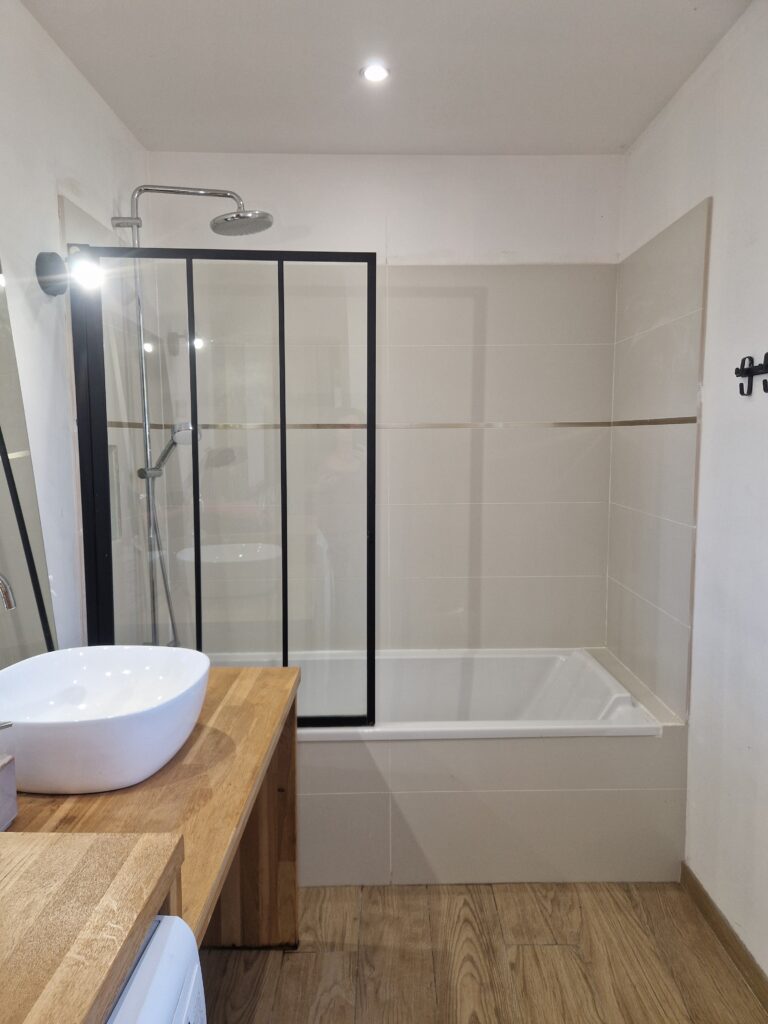 Une salle de bain moderne et lumineuse équipée d'une baignoire avec pare-douche noir et d'une vasque posée sur un meuble en bois