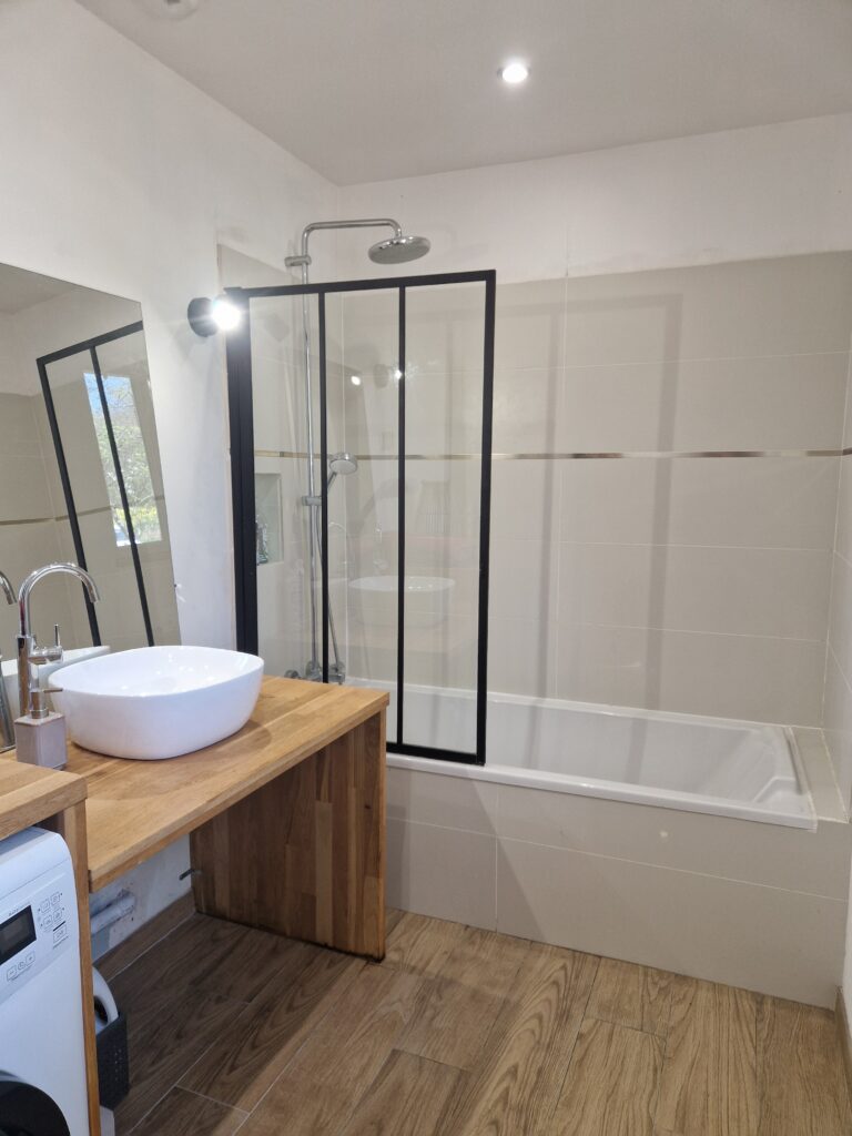 Une salle de bain moderne et lumineuse avec une baignoire-douche, une vasque blanche sur un meuble en bois chaleureux et un sol en parquet.
