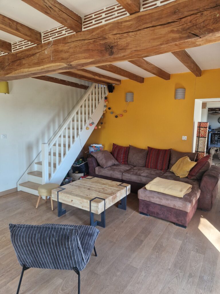 Un salon chaleureux avec poutres apparentes, un grand canapé confortable et un escalier blanc menant à l'étage du gîte.