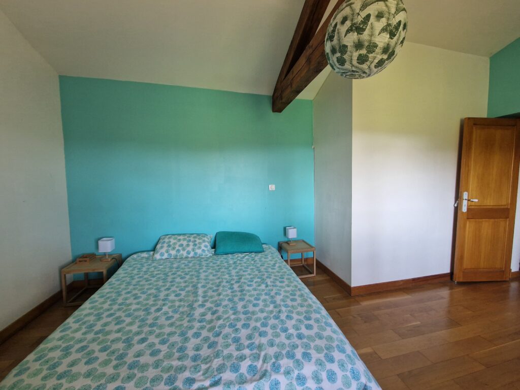 Une chambre lumineuse et chaleureuse avec un lit double, un mur bleu turquoise, des poutres apparentes et un parquet en bois.