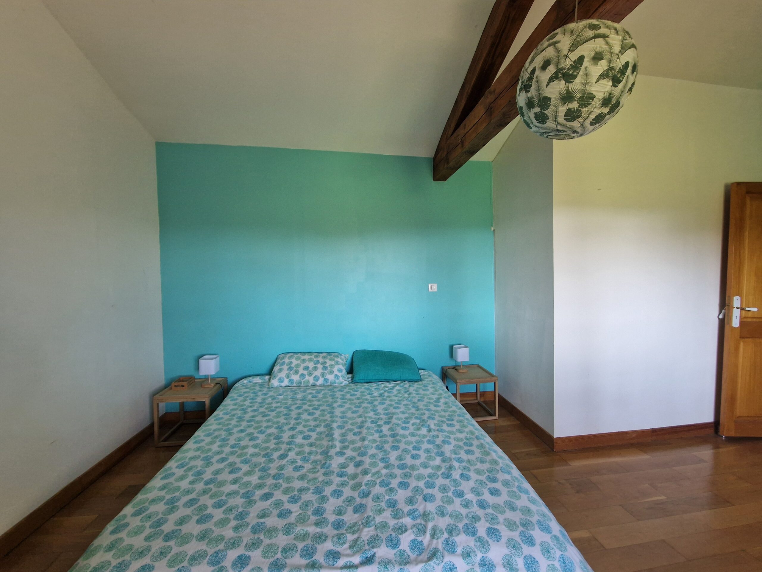 Une chambre lumineuse et chaleureuse avec un mur bleu turquoise, un grand lit confortable et des poutres apparentes en bois.